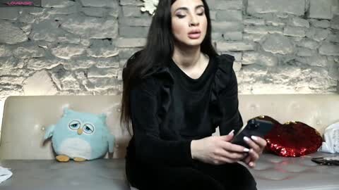 Media: Video of a woman with long dark hair, wearing a black ruffled top, sitting on a beige sofa against a rustic stone wall, holding a smartphone. Background includes a stuffed owl and a red metallic bag.