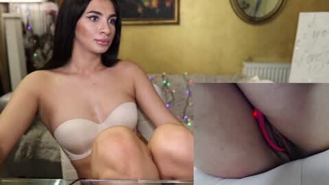 Media: Video of a young woman with long black hair and fair skin, wearing a beige strapless bra, sitting on a bed with a gold wall and Christmas lights in the background.