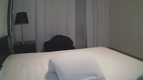 Media: Video of a minimalist hotel room with a white bed, white curtains, a black chair, and a black lamp on a nightstand.