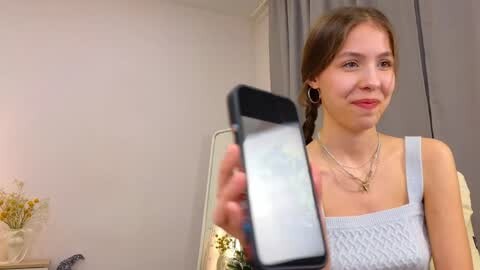 Media: Video of a young Caucasian woman with fair skin and long brown hair in a braid, wearing a light blue patterned dress, holding a smartphone in a bedroom with a mirror and floral decor.