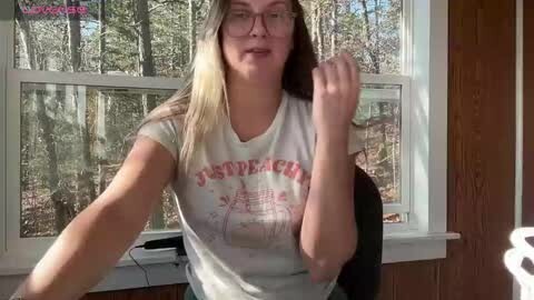 Media: Video of a blonde woman with glasses, wearing a white \"Just Peachy\" t-shirt, seated indoors near a window, surrounded by a forest.