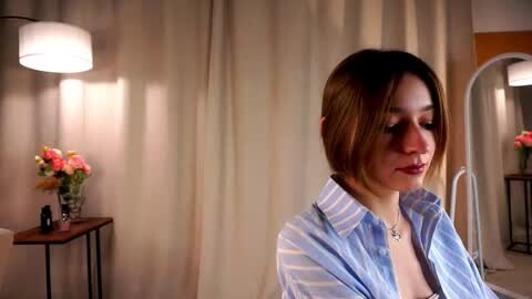 Media: A video of a young woman with light brown hair in a side-parted style, wearing a light blue and white striped shirt. She stands in a warmly lit, softly decorated room with beige curtains and a flower arrangement on a table.