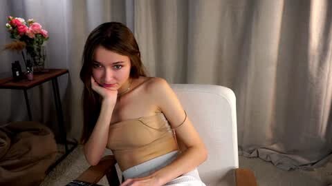 Media: Video of a slender young woman with long brown hair, resting her chin on her hand, wearing a beige strapless top and white pants, seated on a white chair against beige curtains.