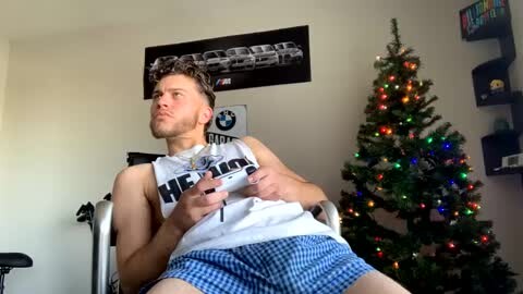 Media: Video of a young man in a white tank top, blue plaid shorts, and headphones, sitting in a wheelchair beside a decorated Christmas tree in a brightly lit room.