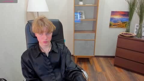 Media: A video of a young man with shoulder-length red hair wearing a black shirt, sitting in a black chair in a room with a wooden dresser, shelves, and a wall with colorful artwork.