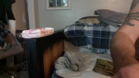 Media: Video of a cluttered bedroom with a black wooden bed, a gray pillow, and a plaid blanket. A person's bare arm with a tattoo is visible.