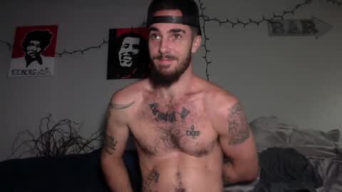 Media: Video of a shirtless, muscular man with a beard and tattoos, wearing a backward baseball cap. Background features two posters and a string of fairy lights.