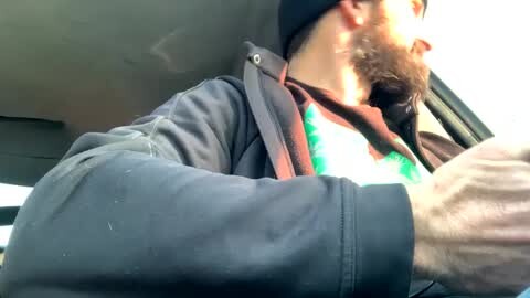 Media: Video of a bearded man wearing a black beanie, gray hoodie, and reflective vest, driving a car with sunlight streaming through the window, creating a dramatic, bright contrast.