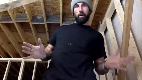Media: Video of a man with a beard, wearing a gray beanie and black t-shirt, hands raised, standing in a wooden-framed room with exposed ceiling joists.
