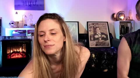 Media: Video of a blonde woman with long hair, topless, smiling, standing near a lit fireplace, surrounded by framed artwork, in a cozy, dimly lit room.