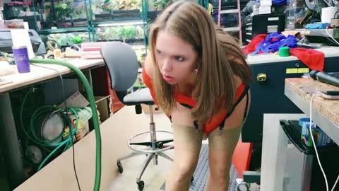 Video of a young Caucasian woman with long, light brown hair, wearing a red top and beige stockings, bending over in a messy aquarium shop. The background features aquariums, tanks, and various fish supplies.