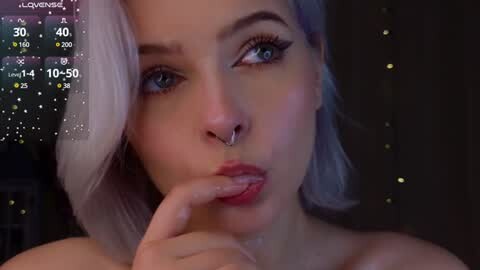 Media: Video of a young, light-skinned woman with platinum blonde hair, blue eyes, and a septum piercing. She's making a seductive face, with her index finger teasing her lips. The background shows a dimly lit room with twinkling fairy lights.