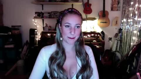 Media: Video of a smiling, fair-skinned woman with long, wavy, light brown hair, wearing a white top, in a dimly lit room with guitars, musical equipment, and string lights.