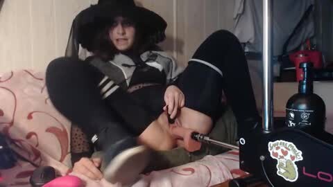 Media: Video of a young woman with pale skin, wearing a witch hat, black lingerie, and thigh-high socks, playing a recorder on a bed with red sheets, surrounded by a red kettle and a bottle of wine.
