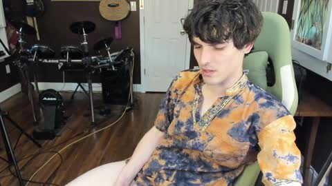 Media: A video of a young, fair-skinned, curly-haired man in a tie-dye shirt, sitting on a green chair in a music studio with equipment, wooden floor, and white doors.