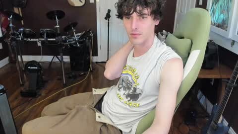 Media: Video of a young, fair-skinned man with curly hair, wearing a white sleeveless shirt and beige pants, sitting in a green gaming chair, surrounded by a drum set and various instruments in a cluttered, dimly lit room.