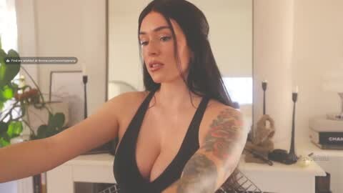 Media: A video of a curvy, light-skinned woman with long black hair, wearing a black halter top, showcasing her large breasts and tattoos on her arm. She stands in a bright, modern bathroom with white walls, a sink, and plants.