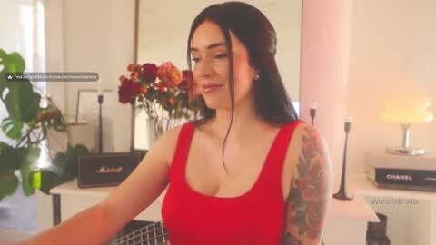 Media: Video of a young woman with long black hair and a tattooed arm in a red tank top, smiling in a modern, well-lit room with a floral arrangement and a lamp on a white counter in the background.