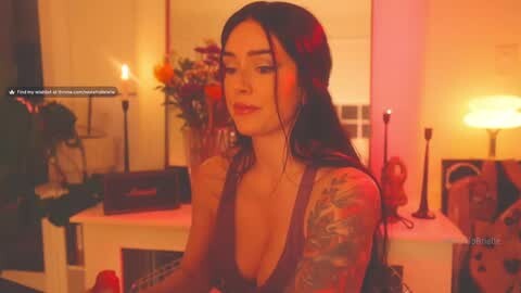 Media: Video of a tattooed woman with long black hair in a low-cut top, sitting in a dimly lit room with candles, flowers, and books.