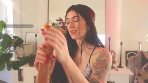 Media: Video of a smiling, tattooed woman with long black hair, wearing a floral bralette, holding a tall glass of brown liquid, in a modern, minimalistic living room with plants and decorative items.