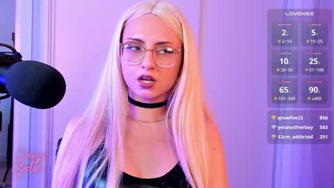 Media: Video of a blonde woman with glasses and a black choker, standing in front of a purple background. Live stream overlay shows viewers and chat.
