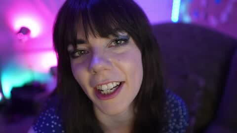 Media: Video of a young woman with straight, dark hair and bangs, smiling broadly with braces. She wears a blue patterned top. Background features blurred, colorful lights in pink and blue.