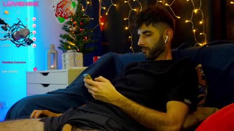 Media: Video of a bearded man in a black t-shirt, sitting on a blue couch, playing a video game with headphones, surrounded by festive decorations and a lit Christmas tree.