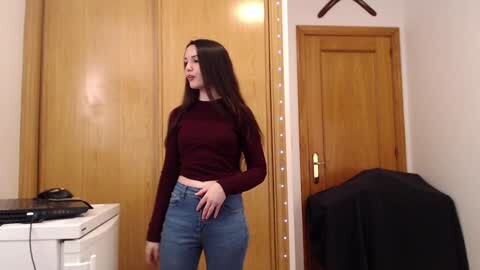 Media: Video of a young woman with long dark hair, wearing a maroon long-sleeve top and high-waisted blue jeans, standing in a cozy, wood-paneled room with a closed door, a white appliance, and a black garment draped over a chair.