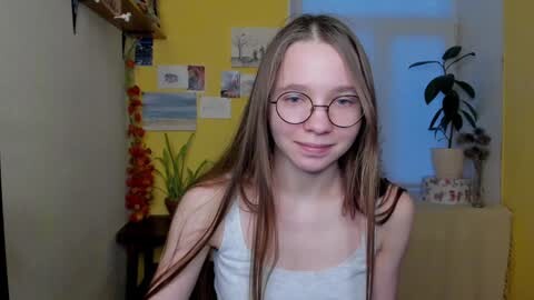 Media: Video of a young Caucasian woman with long, straight light brown hair, wearing glasses, a white tank top, and smiling in a cozy, yellow-painted room with potted plants, artwork, and a red orchid.
