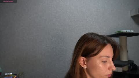 Media: Video of a woman with long brown hair, wearing earrings, standing against a gray textured wall. Background includes a black shelf with a white vase and a green plant, and a beige cat tree.