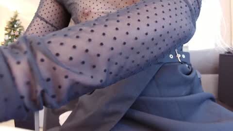 Media: Video of a woman in sheer, polka-dotted blouse, blue jeans, and white underwear, sitting on a sofa. Background shows a Christmas tree.