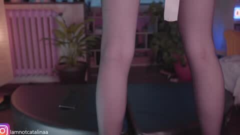 Media: Video of a woman's legs in pink leggings, standing on a bed in a dimly lit room with potted plants and a radiator in the background.