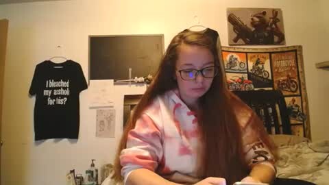 Media: Video of a young woman with long brown hair, wearing glasses and a tie-dye pink shirt, sitting in a cozy room with a Star Wars poster, black shirt on wall, and a flat-screen TV.