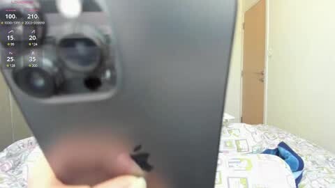 Media: Video of a person's hand holding a gray iPhone, displaying camera settings. Background features a bed with a white and green patterned blanket and a partially visible wooden door.