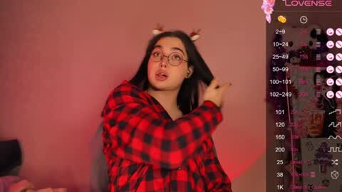 Media: Video of a young woman with long black hair, wearing glasses and a red plaid shirt, playfully touching her hair against a pink wall.