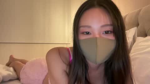Media: Video of an Asian woman with long black hair, wearing a pink bra, lying on a bed with a beige headboard and fluffy pink pillow.