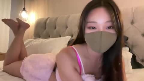 Media: Video of an Asian woman in pink lingerie and face mask lying on a bed, head turned, feet playfully raised.