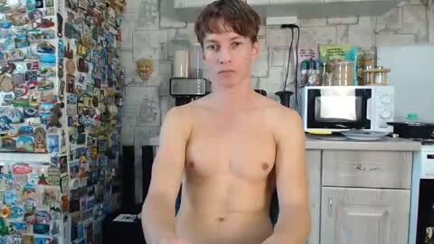 Media: Video of a shirtless, fair-skinned, short-haired young man with small breasts, standing in a cluttered, colorful kitchen with a microwave, stove, and many small appliances.