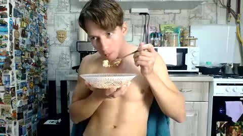 Media: A video of a shirtless, light-skinned, slender young man with short brown hair eating from a white bowl in a cluttered, colorful kitchen.