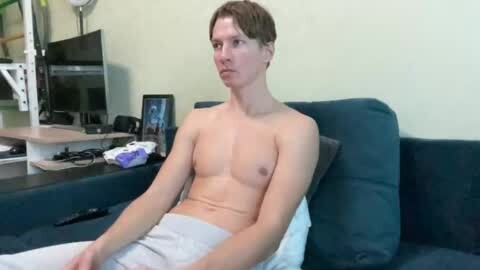 Media: Video of a young, fair-skinned, shirtless man with short brown hair, sitting on a blue couch, wearing light pajama pants, in a room with a TV, a computer, and a bookshelf.