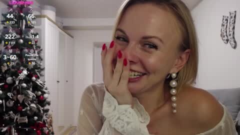 Media: Video of a smiling, fair-skinned blonde woman with straight hair, wearing pearl earrings and a sheer white lace top, posing indoors with a decorated Christmas tree in the background.