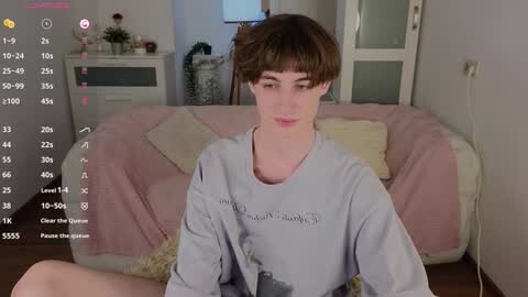 Media: Video of a young, fair-skinned boy with short, light brown hair in a grey graphic tee, sitting on a pink couch in a cozy, minimalist room.