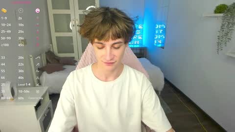 Media: Video of a young man with short brown hair, wearing a white T-shirt, sitting in a modern bedroom with a bed, blue light, and digital clock.