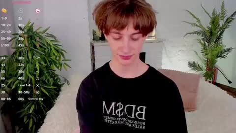 Media: A video of a young, pale-skinned, androgynous person with short brown hair wearing a black \"BAM\" t-shirt, smiling softly. Background features a green potted plant, a furry blanket, and a white wall.
