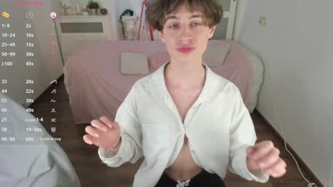Media: Video of a young, androgynous person with short, brown hair, wearing a white button-up shirt, sitting in a pink draped room.