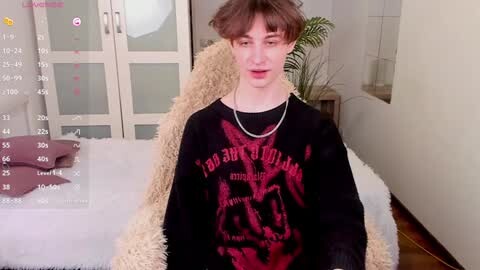 Media: Video of a young, slim, pale-skinned boy with messy brown hair, wearing a black sweatshirt with a red devil graphic, sitting on a beige couch in a modern, minimalist living room.