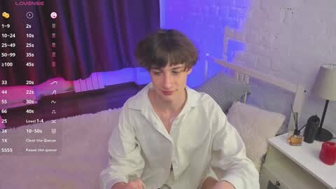 Media: Video of a young, Caucasian male with short brown hair, wearing a white button-up shirt, seated on a bed in a dimly lit bedroom with purple lighting.