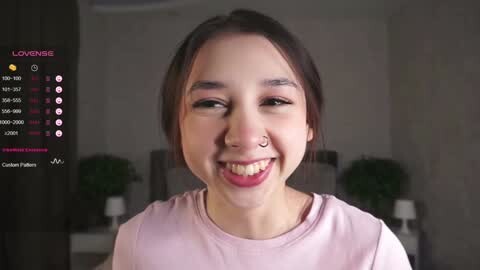 Media: Video of a smiling Asian woman with dark hair, wearing a pink shirt, and a nose ring, set in a dimly lit room with greenery and a TV displaying streaming stats.