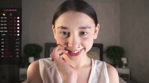 Media: Video of a smiling Asian woman with light skin and straight dark hair, wearing a white sleeveless top, in a dimly lit room with a calendar on the left.