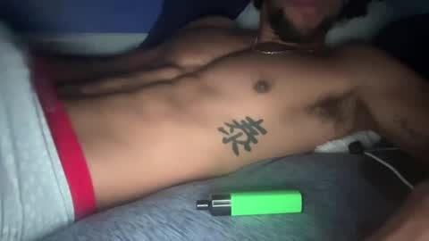 Media: Video of a shirtless man with a toned, tan body and a tattoo on his chest. He's lying on a bed, wearing white pants with a red waistband. A green and black vape pen lies beside him.